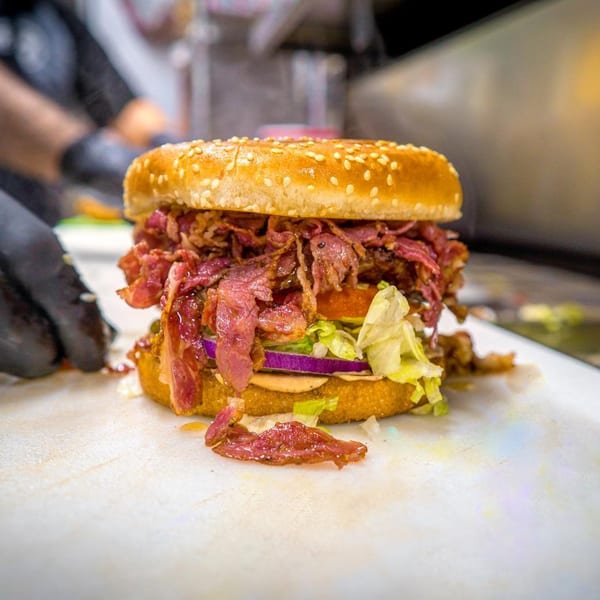 A Frosty King burger with pastrami, lettuce, tomato, and red onion on a sesame seed bun.
