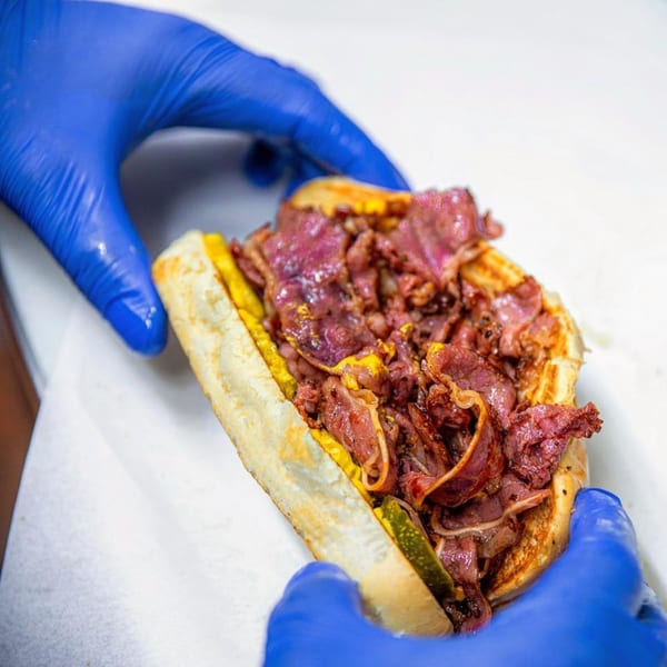 Gloved hands holding a Frosty King burger with pastrami, mustard, and pickles on a bun.