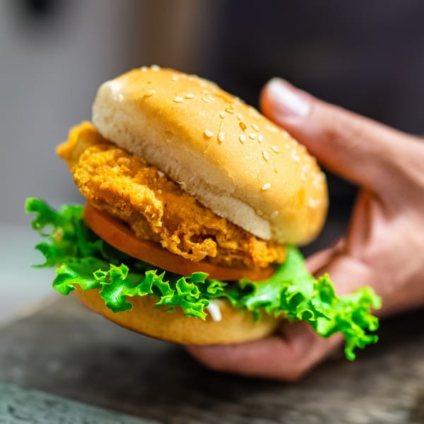 Hand holding a Frosty King crispy chicken burger with lettuce, tomato, and sesame seed bun.