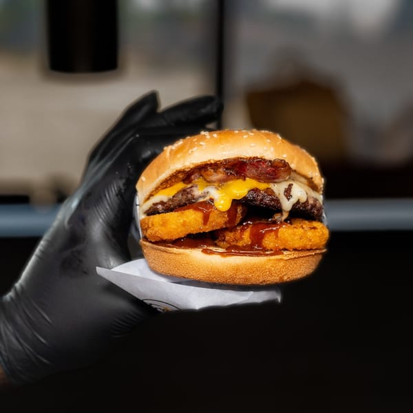 A gloved hand holds a Frosty King burger with hash browns, cheese, bacon, and BBQ sauce.