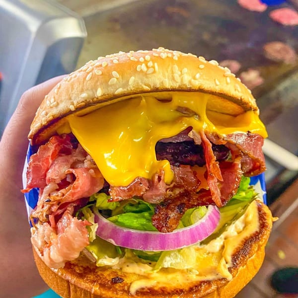A hand holds a Frosty King bacon cheeseburger with melted cheese, lettuce, and red onion.